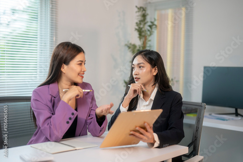 Two women are sitting at a desk, one of them holding a clipboard