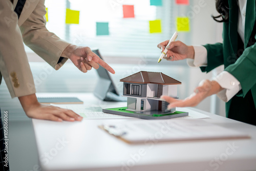 Two women are working on a house model, one pointing out a flaw