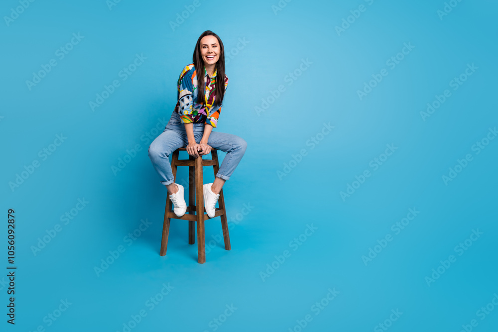 Full length photo of pretty nice pleasant woman wear print shirt denim trousers sitting on bar stool isolated on blue color background Full length photo of pretty nice pleasant woman wear print shirt denim trousers sitting on bar stool isolated on blue color background