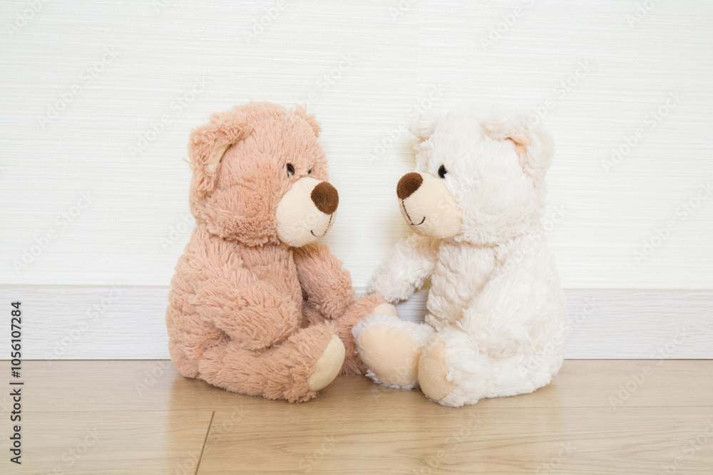 Two white and light brown soft teddy bears looking at each other and sitting on wooden floor at light gray wall background. Closeup. Children communication concept. Front view. Two white and light brown soft teddy bears looking at each other and sitting on wooden floor at light gray wall background. Closeup. Children communication concept. Front view.