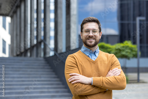 Close-up portrait of a young smiling male businessman standing outside an office center wearing a sweater and glasses, crossing his arms over his chest and looking sullenly at the camera photo