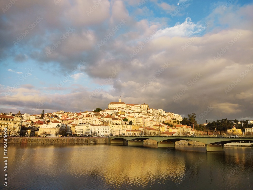 Fototapeta premium This image captures a scenic view of a historical city built on a hillside, likely Coimbra, Portugal, during a picturesque moment with the late afternoon or early evening sunlight