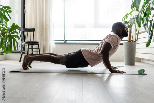 Young African-American man doing yoga on a mat in a bright home setting, practicing the cobra pose with focused posture. Surrounded by plants, promoting wellness and relaxation.