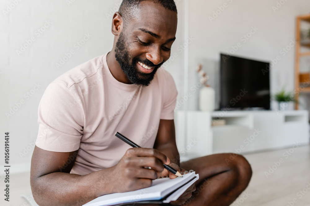 Young African-American man sitting cross-legged on a yoga mat at home, writing in a notebook after a workout, with a smartphone nearby, creating a calm and focused atmosphere. Young African-American man sitting cross-legged on a yoga mat at home, writing in a notebook after a workout, with a smartphone nearby, creating a calm and focused atmosphere.