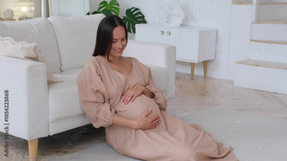 Smiley pregnant middle aged Caucasian woman gently smoothing a baby bump sitting on floor at home living room. Concept of a healthy pregnancy and wellness. Smiley pregnant middle aged Caucasian woman gently smoothing a baby bump sitting on floor at home living room. Concept of a healthy pregnancy and wellness.