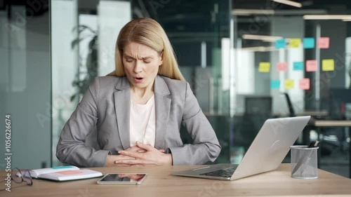 Sick businesswoman in jacket suffering from stomach pain while working on laptop sitting at workplace in business office. Upset female employee has gastritis, poisoning, bloating or digestive problems