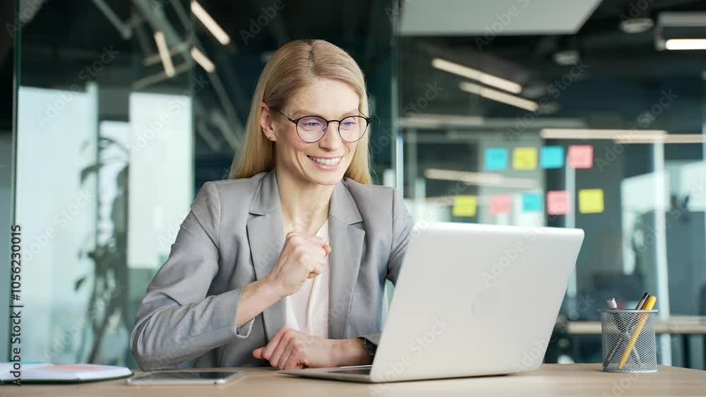 Satisfied happy businesswoman in jacket reading great news on laptop while sitting at desk at workplace in business office. Smiling glad woman smiles, rejoices at a positive message on the computer Satisfied happy businesswoman in jacket reading great news on laptop while sitting at desk at workplace in business office. Smiling glad woman smiles, rejoices at a positive message on the computer