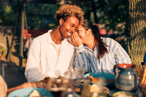 Woman whispering in male friend's ear sitting at dining table during dinner party photo