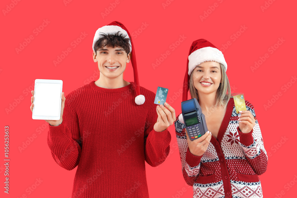 Happy young couple in Santa hats with tablet computer, credit cards and payment terminal on red background. Online shopping for Christmas