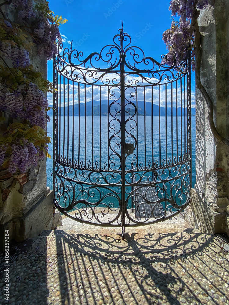 View of Lake Como and mountains through closed iron gate, Italy against blue sky with clouds