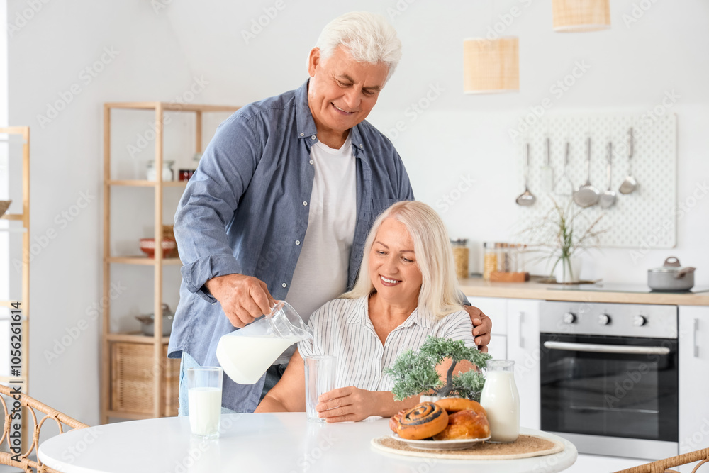 Happy senior couple with tasty buns pouring milk from jug into glasses in kitchen Happy senior couple with tasty buns pouring milk from jug into glasses in kitchen