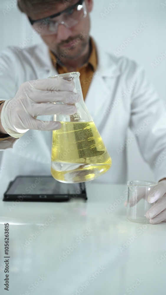 Man scientist wearing white protective gloves, and glasses is holding a yellow chemical solution inside an Erlenmeyer flask in a laboratory, vertical portrait. Science and medicine Man scientist wearing white protective gloves, and glasses is holding a yellow chemical solution inside an Erlenmeyer flask in a laboratory, vertical portrait. Science and medicine