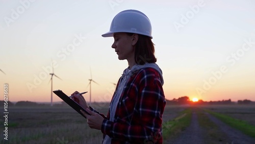 Woman engineer is taking notes on a clipboard on a field with wind turbines, as the sun sets in evening. Clean energy and engineering