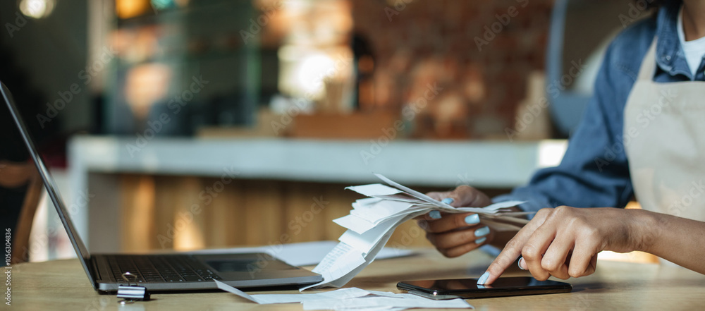 Payment of bills, bankruptcy and modern cafe during covid-19 quarantine. Millennial african american woman bartender in apron sits at table and works with laptop in interior of cafeteria, cropped Payment of bills, bankruptcy and modern cafe during covid-19 quarantine. Millennial african american woman bartender in apron sits at table and works with laptop in interior of cafeteria, cropped