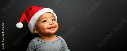 Adorable African American little baby in Santa hat smiling and looking up on black banner. Curious baby dressed for holiday celebrations. photo