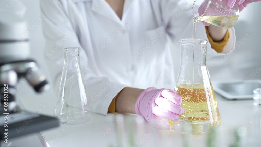 A scientist, wearing a lab coat and pink protective gloves, is pouring a yellow oily liquid from one beaker to another near microscope in laboratory, close up. Medicine and science A scientist, wearing a lab coat and pink protective gloves, is pouring a yellow oily liquid from one beaker to another near microscope in laboratory, close up. Medicine and science