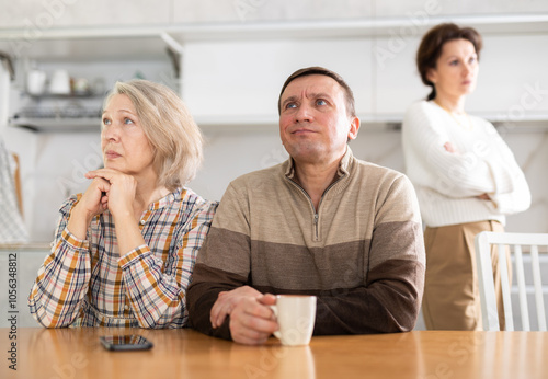 Livid middle-aged man sitting at the kitchen-table next to old female family member looking upset