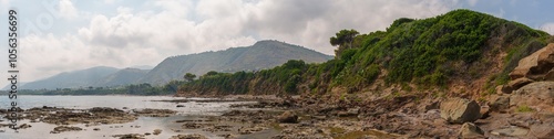 A wide-angle panoramic view of a rugged coastal landscape featuring rocky shores, lush green cliffs, and distant hills under a partly cloudy sky. Ideal for nature and travel photography