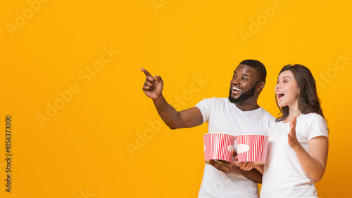 Cinema Offer. Excited interracial couple holding popcorn backets and pointing aside at copy space over yellow background, panorama