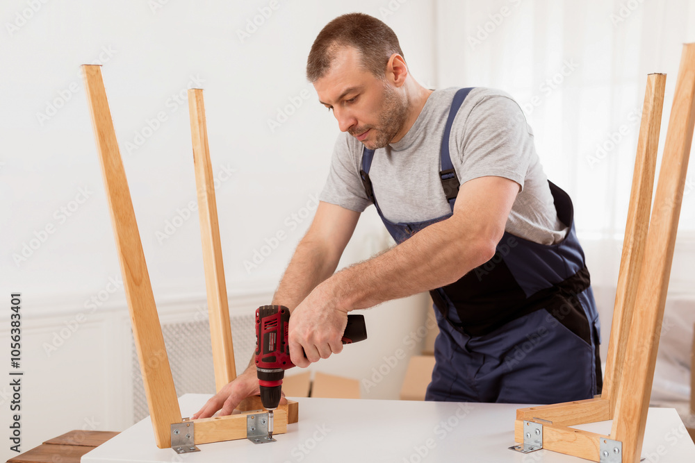 Male Carpenter Using Electric Drill Assembling Wooden Table After Transportation To New Apartment. Man In Blue Coverall Uniform Fixing Desk With Carpentry Tools Working In Studio. Male Carpenter Using Electric Drill Assembling Wooden Table After Transportation To New Apartment. Man In Blue Coverall Uniform Fixing Desk With Carpentry Tools Working In Studio.