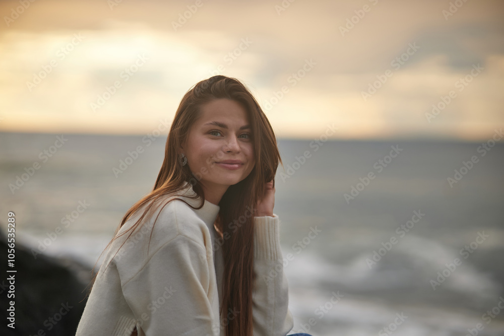 A young woman sits on a rocky shore, gazing into the distance with a calm expression. Dressed casually in jeans and a light sweater, she enjoys the serene ocean backdrop at sunset. A young woman sits on a rocky shore, gazing into the distance with a calm expression. Dressed casually in jeans and a light sweater, she enjoys the serene ocean backdrop at sunset.