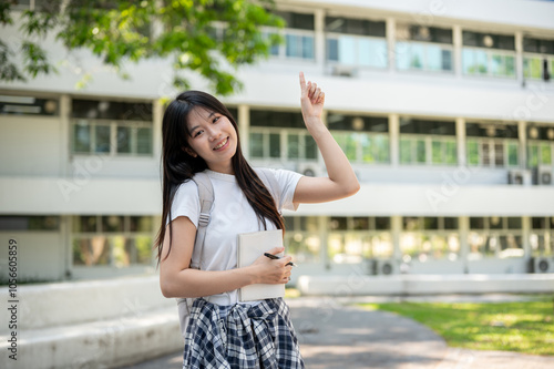 A charming Asian female student standing on her college campus, holding a book, raising one finger.