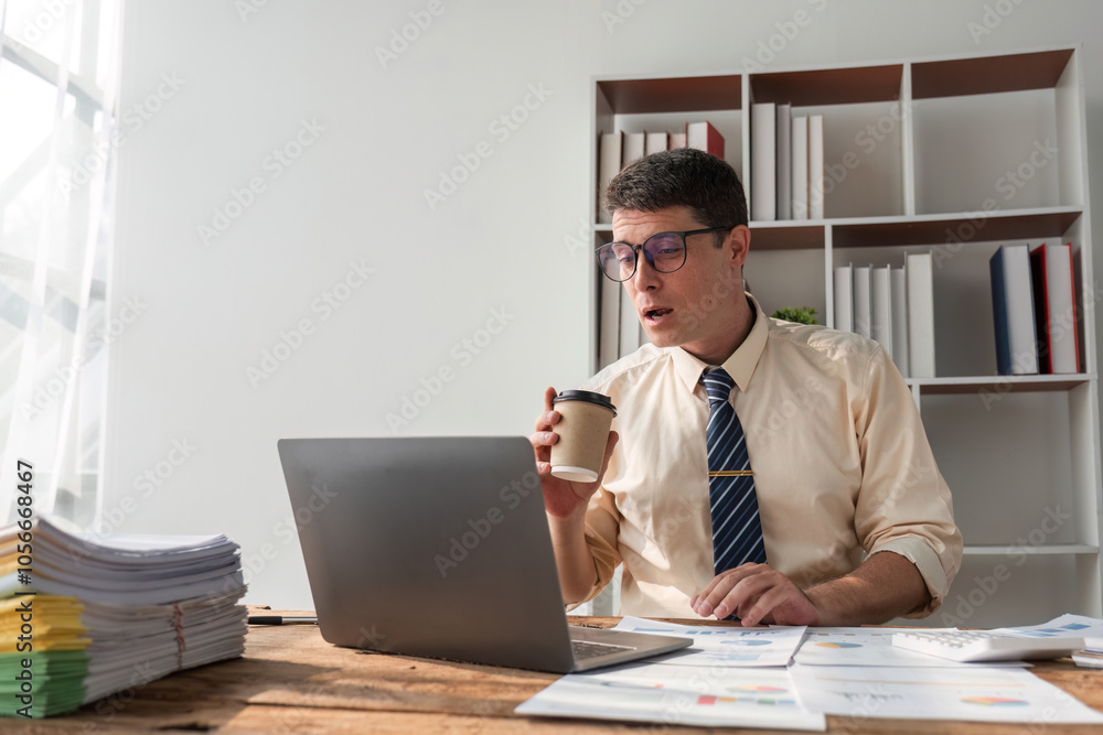Young Businessman Working in Modern Office with Laptop and Coffee, Analyzing Documents and Data Young Businessman Working in Modern Office with Laptop and Coffee, Analyzing Documents and Data