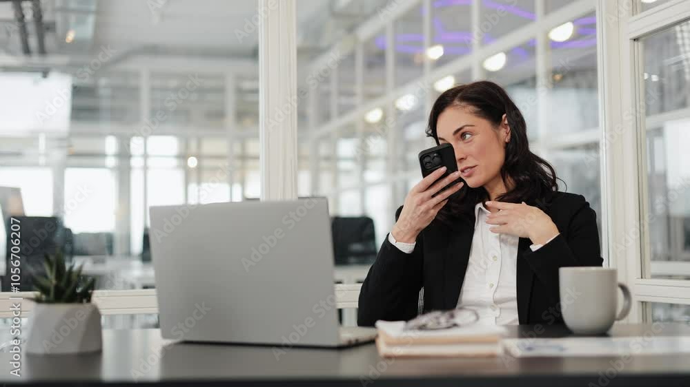 Positive businesswoman in a suit sits at her desk in office, talking on speakerphone, gesturing. Laptop is open in front of her Modern leader person explaining details to business partner at workplace Positive businesswoman in a suit sits at her desk in office, talking on speakerphone, gesturing. Laptop is open in front of her Modern leader person explaining details to business partner at workplace