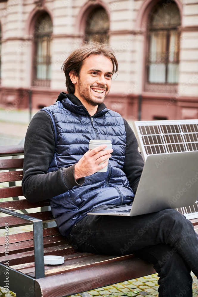 Man sits on wooden bench in urban setting, working on laptop with cup of coffee in hand. Solar panel rests beside, emphasizing sustainable, mobile workspace amidst backdrop of historic architecture. Man sits on wooden bench in urban setting, working on laptop with cup of coffee in hand. Solar panel rests beside, emphasizing sustainable, mobile workspace amidst backdrop of historic architecture.