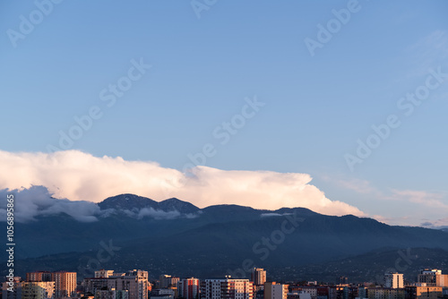 Cityscape of panel buildings in warm orange sunset light, framed by distant mountains and a clear blue sky, capturing the peaceful contrast of urban life against a natural backdrop