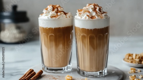 Two tall glasses of warm coffee topped with whipped milk foam cinnamon and caramel on a marble surface