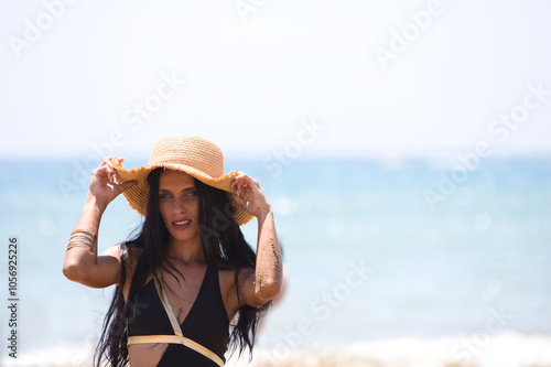 Young and beautiful brunette woman with green eyes is on the beach in swimsuit and puts a straw hat on her head and holds it with her hands. In the background the horizon with the sea and blue sky.