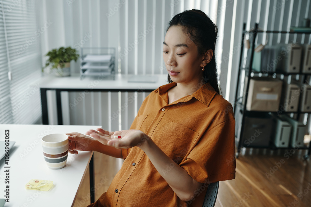Asian pregnant woman relaxing with cup sitting at an office desk. She appears thoughtful while holding cup, surrounded by modern office furnishings and natural light Asian pregnant woman relaxing with cup sitting at an office desk. She appears thoughtful while holding cup, surrounded by modern office furnishings and natural light