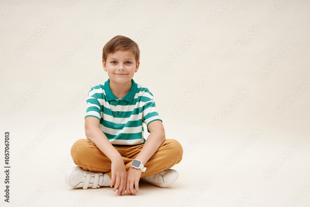Cute little boy wearing vibrant striped polo t shirt sitting cross legged while smiling at camera on neutral background in studio, copy space Cute little boy wearing vibrant striped polo t shirt sitting cross legged while smiling at camera on neutral background in studio, copy space