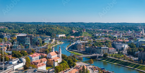 Aerial view of Vilnius, Lithuania, featuring Gediminas' Tower on a green hill, the Neris River, and a mix of modern and traditional architecture.