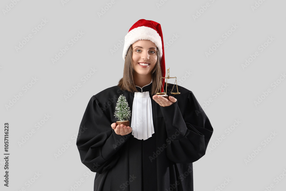 Female judge in Santa hat with justice scales and toy Christmas tree on light background Female judge in Santa hat with justice scales and toy Christmas tree on light background
