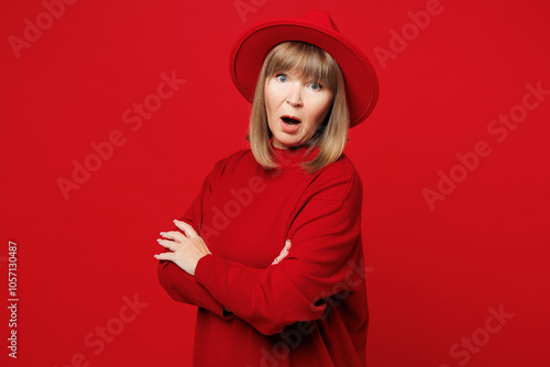 Side profile view sad elderly blonde woman 50s years old wearing sweater hat casual clothes hold hands crossed folded look camera isolated on plain red background studio portrait. Lifestyle concept.