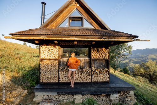 Woman in an orange sweater standing in front of a rustic cabin with stacked firewood, surrounded by a scenic hillside landscape during sunset