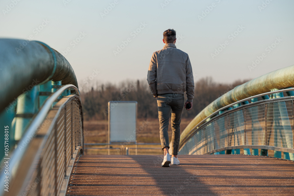 giovane uomo adulto italiano, ritratto di spalle, a figura intera, vestito con abiti casual moderni, mentre cammina su un ponte, di pomeriggio