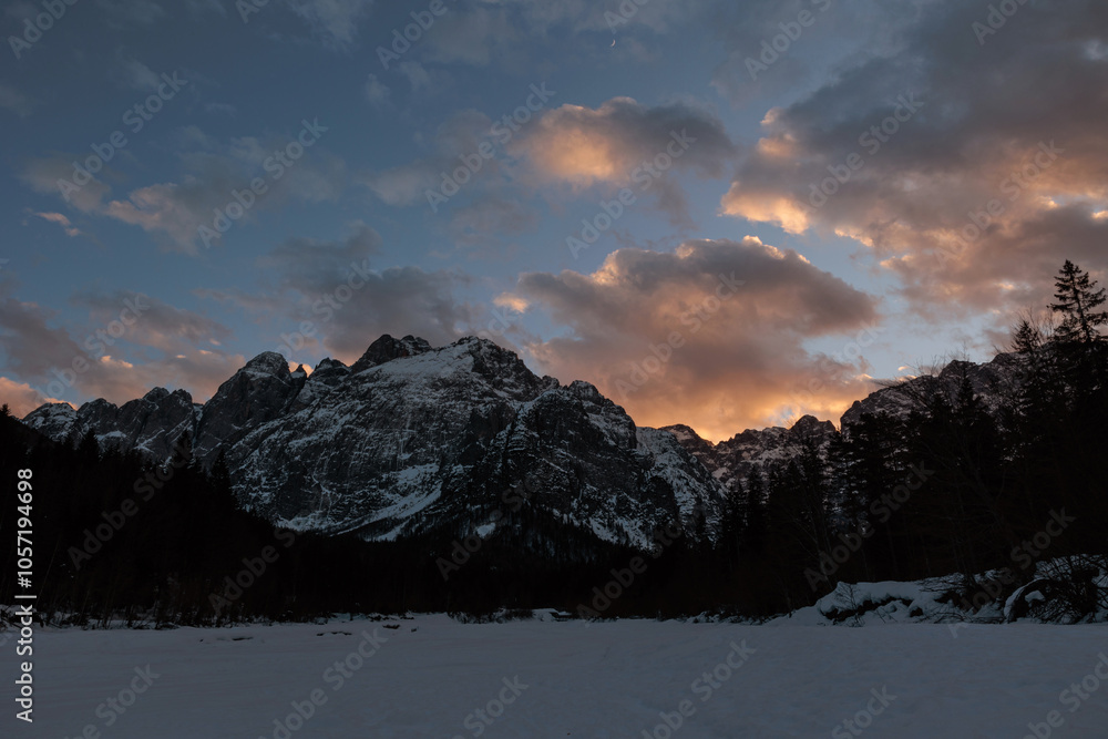 sole che tramonta dietro a delle alte montagne innevate, in un cielo azzurro e parzialmente nuvoloso, con nuvole colorate di arancione, nelle Alpi Italiane, in inverno
