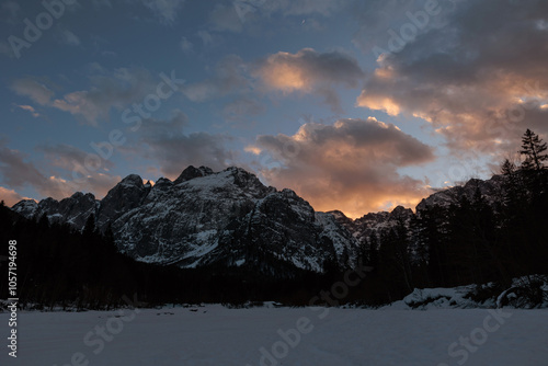 Obraz na płótnie sole che tramonta dietro a delle alte montagne innevate, in un cielo azzurro e parzialmente nuvoloso, con nuvole colorate di arancione, nelle Alpi Italiane, in inverno w salonie