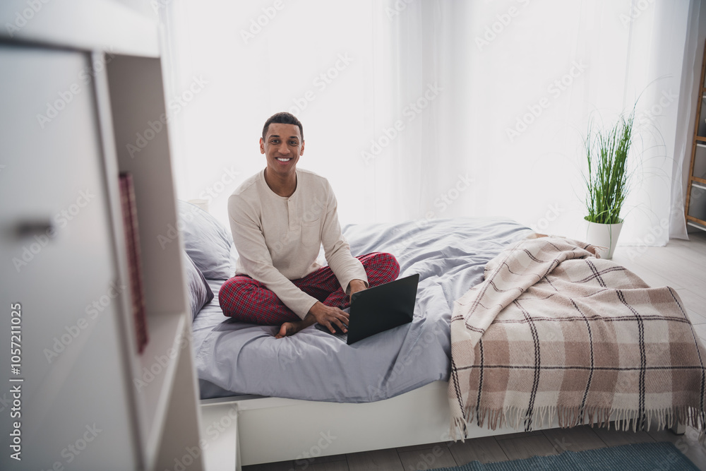 Full length photo of handsome young guy sit bed working netbook dressed sleepwear wake up spacious house indoors bedroom Full length photo of handsome young guy sit bed working netbook dressed sleepwear wake up spacious house indoors bedroom