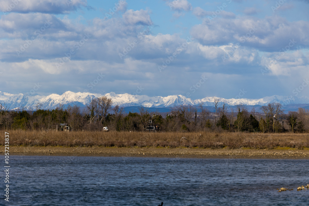 ambiente naturale nel nord Italia, che si estende da un'area di palude fluviale sul mare fino alle catene montuose innevate lontane, con cielo nuvoloso, in inverno