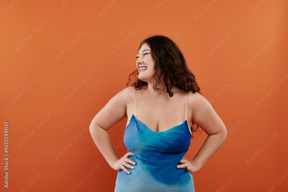 A young plus size woman showcases her style in a blue dress, smiling joyfully with an orange backdrop. A young plus size woman showcases her style in a blue dress, smiling joyfully with an orange backdrop.