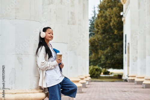 A young Asian student with headphones smiles while leaning against a column, holding coffee and books on campus.