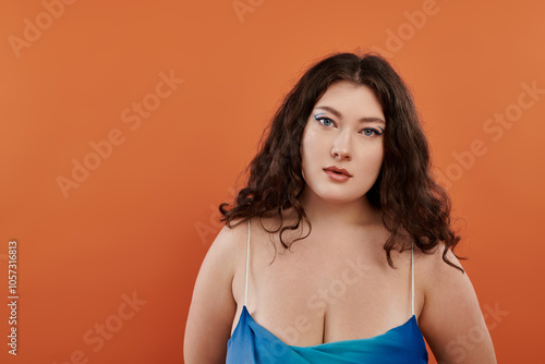 A confident plus size woman showcases her beauty with striking makeup and a flowing blue dress on a bold backdrop.