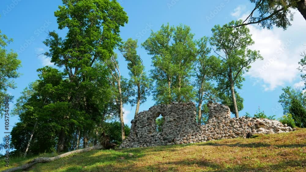 Ancient Stone Walls of Helme Castle Ruins Against Summer Sky in Estonia. Historic Medieval Fortress Remains Surrounded by Tall Green Trees on Hillside Ancient Stone Walls of Helme Castle Ruins Against Summer Sky in Estonia. Historic Medieval Fortress Remains Surrounded by Tall Green Trees on Hillside