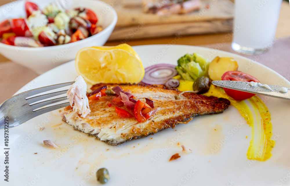 Grilled sea bass in a white porcelain plate. Garnish: lemon, onion, tomato, capers, broccoli, potatoes, carrots.