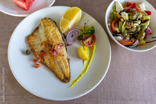 Obraz na płótnie Grilled sea bass in a white porcelain plate. Garnish: lemon, onion, tomato, capers, broccoli, potatoes, carrots. w salonie