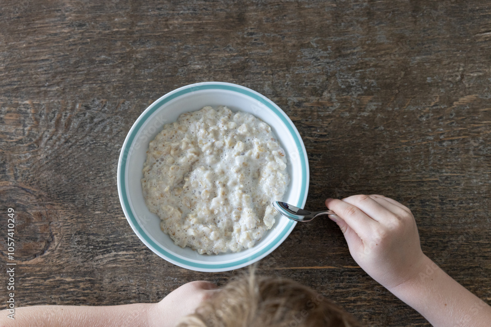 Freshly cooked oatmeal in a white porcelain bowl with blue rim on a rustic wooden dining table. A young light haired boy is sitting at the table holding a spoon about to start eating.